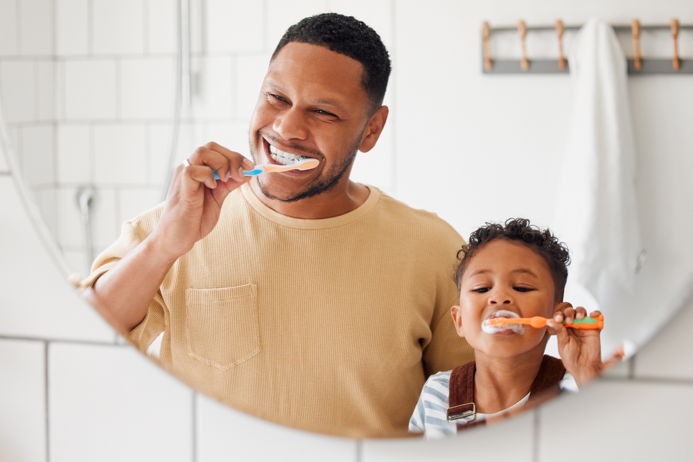 Brushing teeth, father and child in a home bathroom for dental health and wellness with smile. Face of a man and african boy kid learning to clean mouth with a toothbrush and mirror for oral hygiene
